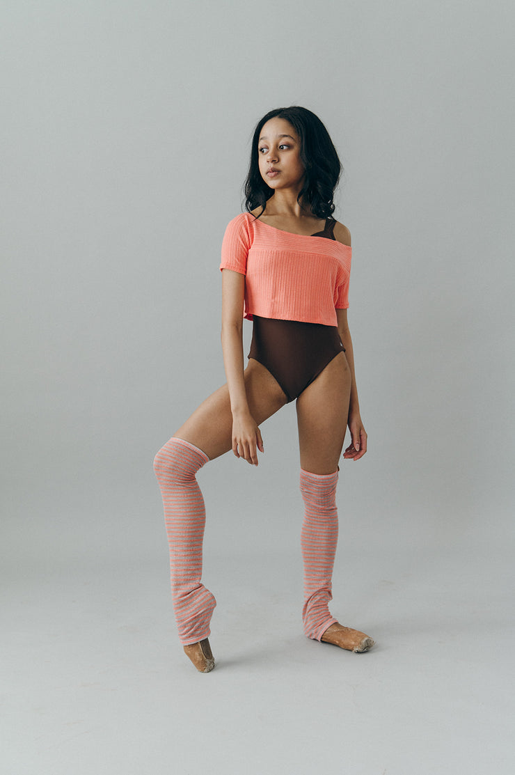 Person wearing a coral top and brown leotard with pink striped legwarmers on a gray background