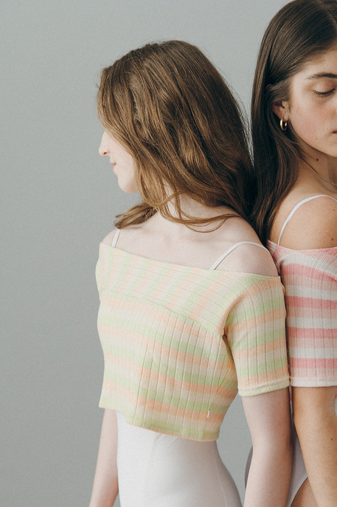 Two women wearing colorful striped tops and white bottoms against a gray background
