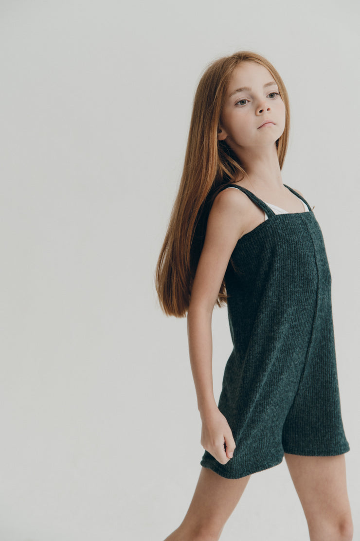 Young girl wearing a green sleeveless romper with straps against a plain background